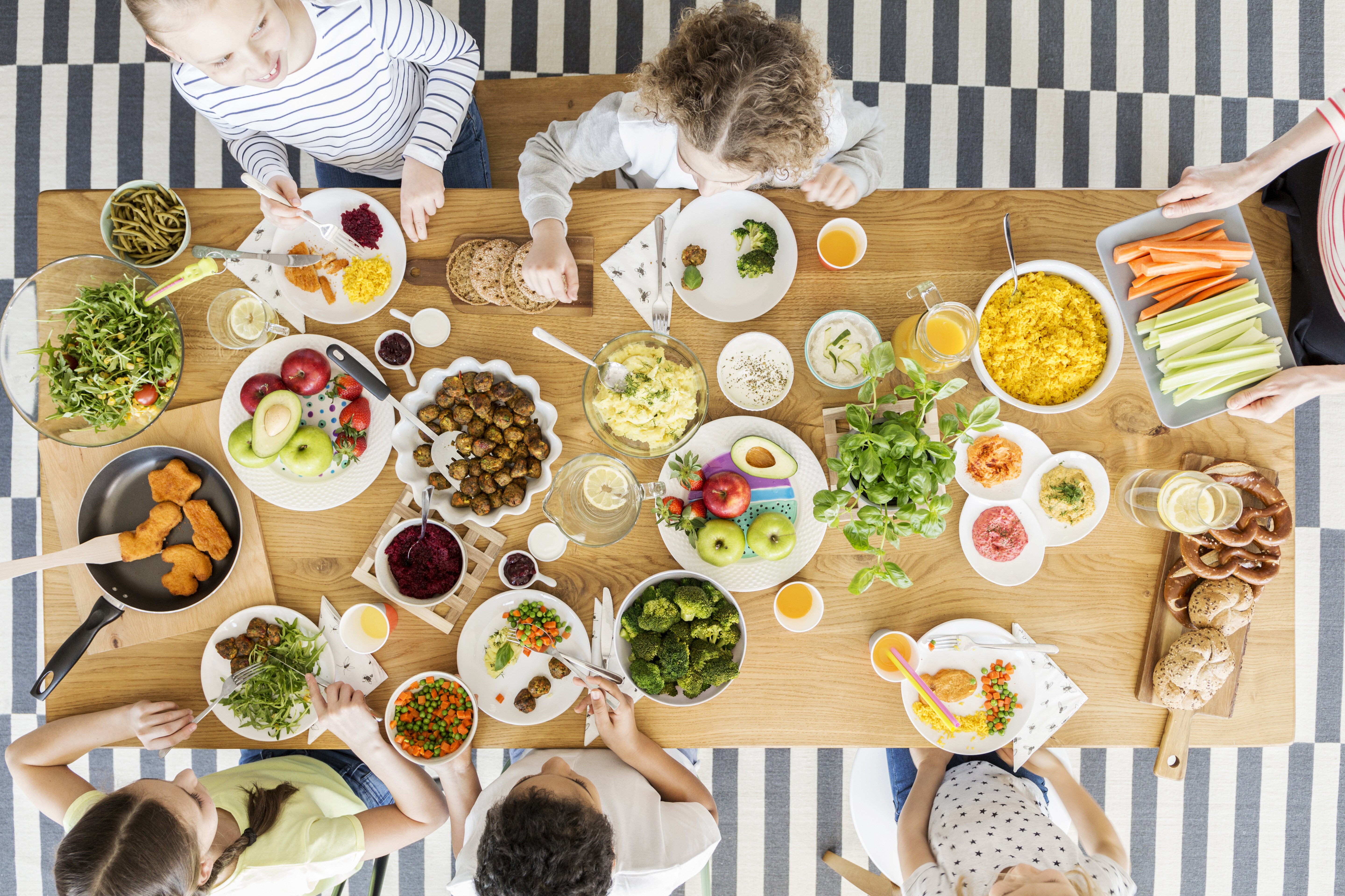 Et stort spisebord set oppefra med masser af frugt og grønt på tallerkner med en lille håndfuld børn i gang med at tage for sig